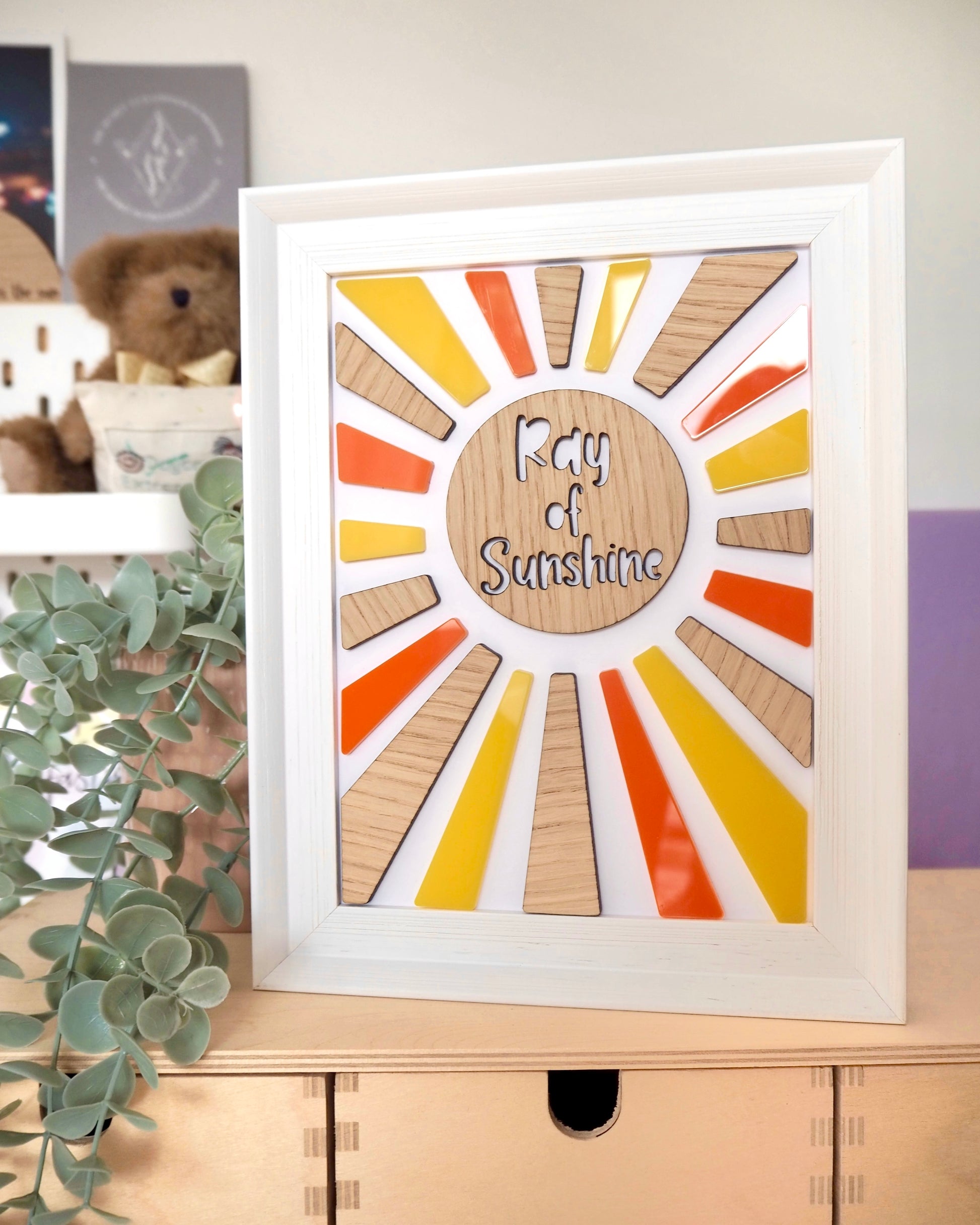 Sunshine made from wood and yellow and orange acrylic for the rays, mounted in a white wooden frame. The words Ray of Sunshine cut out from the middle of the sunshine