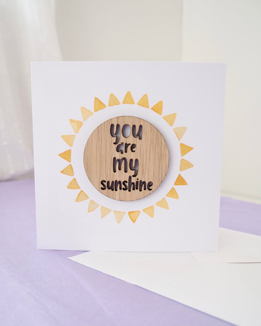 square greetings card with a sunshine which is partly painted in watercolour for the rays and the centre is a circular wooden token that says you are my sunshine