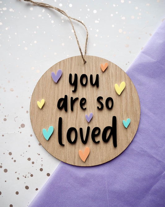 Circle wooden plaque with you are so loved in black acrylic and pastel coloured hearts